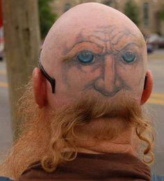 Back of the head of a nearly bald person with a tattoo depicting an expressive face with blue eyes on the back of their head. Their hair is trimmed into a long, luxuriant mustache beneath the tattoo of a nose.