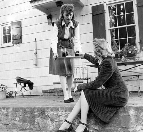 Vintage black and white photo of a woman serving a drink from a tray to another woman seated on a step outside a house. The house appears to be American suburban, the women are well-dressed in middle-American style, the period seems to be 1940s.