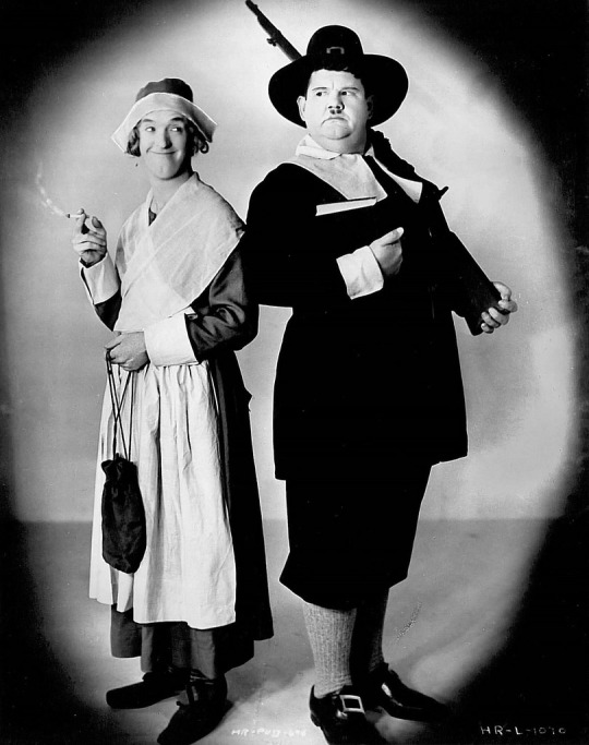 Black and white photo of Laurel and Hardy dressed up as pilgrims. Laurel is carrying a long gun and looking stern, with the pilgrim's characteristic tall hat, breeches and buckle shoes. Laurel is smiling and dressed as a pilgrim woman.