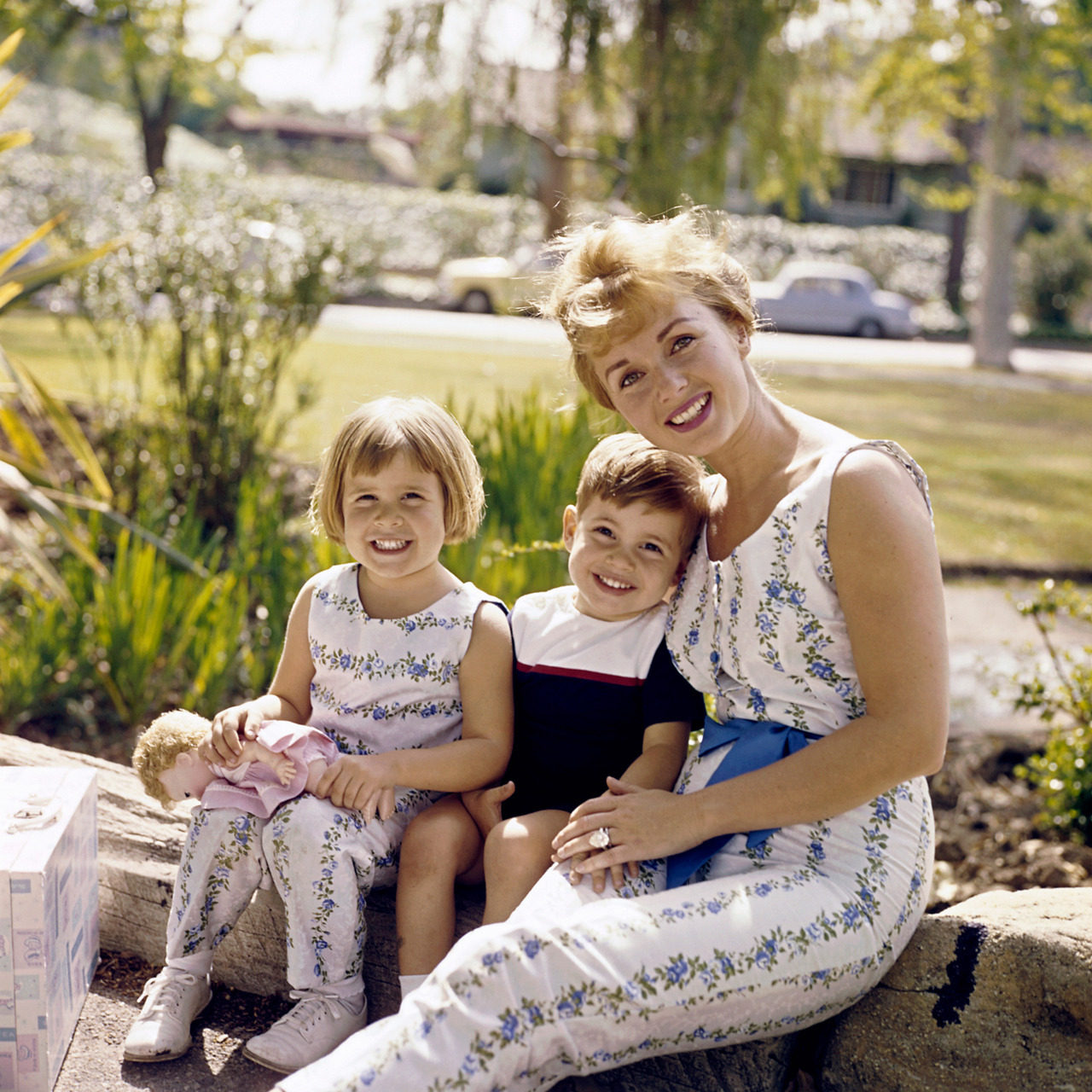 Young, blonde, pretty and wholesome Debbie and her two wholesome and beautiful toddlers are sitting together outdoors, with the little girl holding a doll. Debbie is wearing a kind of floral linen jumpsuit with bare shoulders. Very fetching!