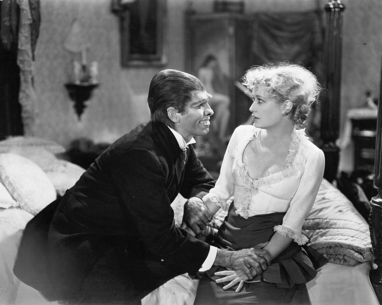 First image in a sequence: Black-and-white movie still of a man in a suit is kneeling beside a woman seated on a bed, engaging in conversation in a vintage setting. She's wearing a bustier. The man is wearing realistic makeup that makes him vaguely resemble a wolf-man.