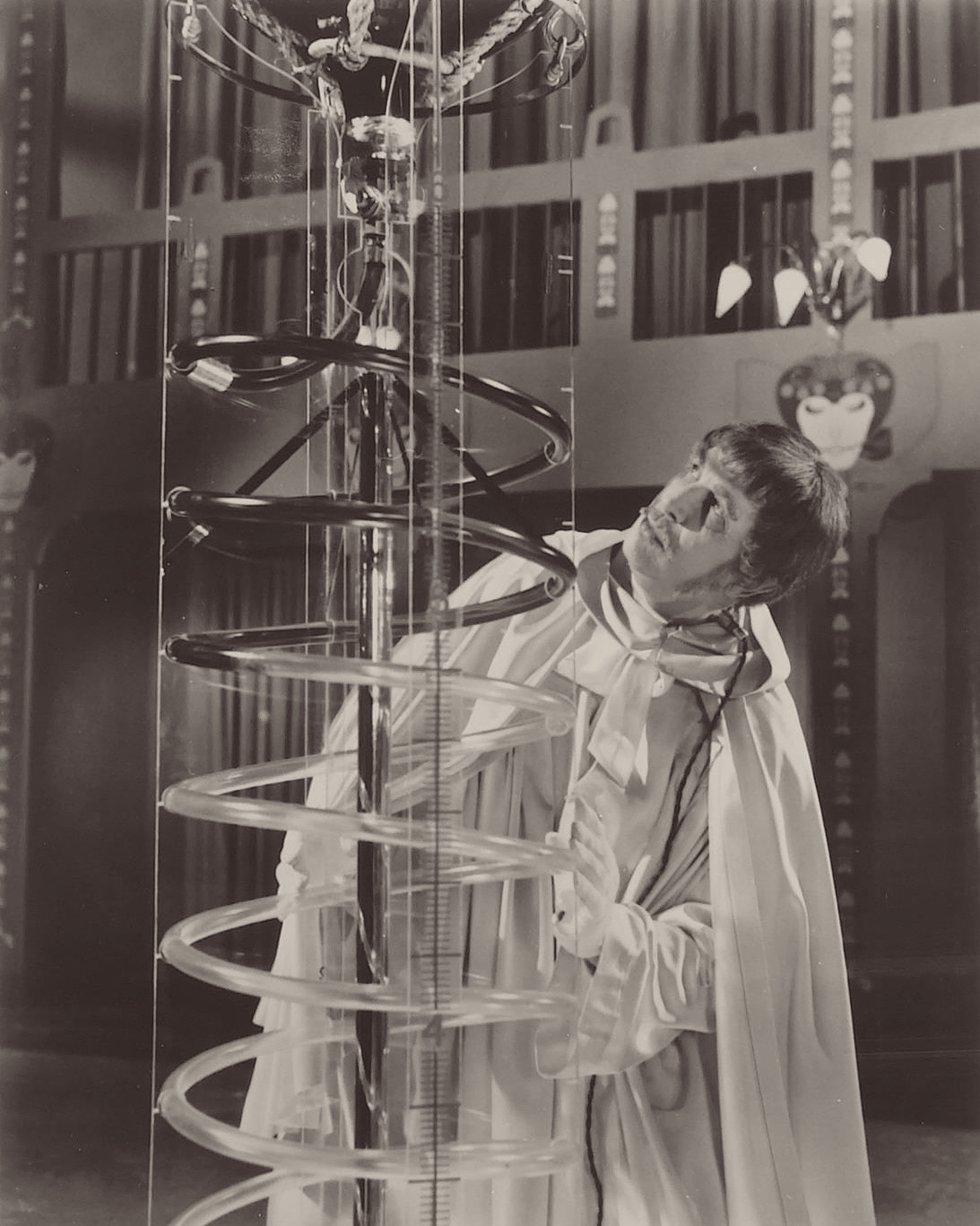 Campy black-and-white movie still of Price standing, wearing a long white robe, kind of a 1966 Beatles haircut and 70s mustache, solemnly examining a large spiral glass apparatus taller than he is, in a dramatic, theatrical setting.