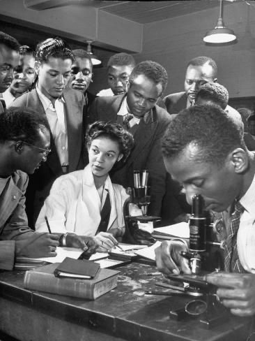 Vintage black-and-white photo, apparently from the 1950s or 1960s, of a group of young Black men and women gather around a laboratory table, watching a Black man  operate a microscope in the foreground. Everyone is neatly groomed; most of the men seem to be wearing neckties.