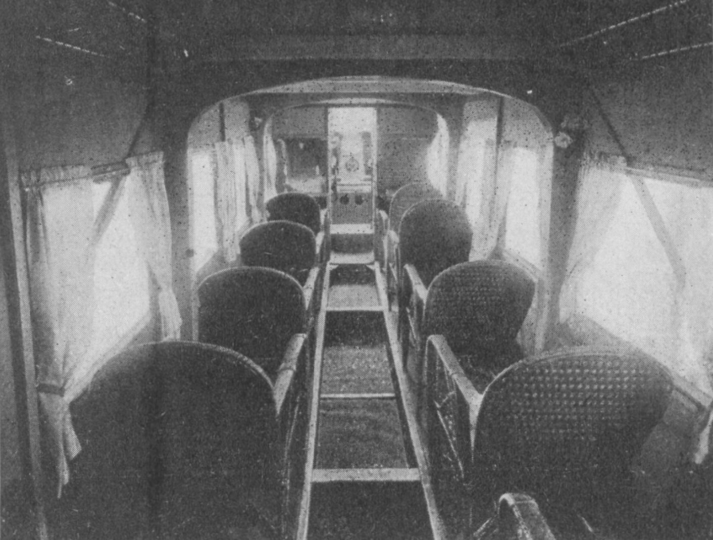 An dark, grainy black-and-white vintage photo of the interior view of an early passenger airplane cabin, shot from the rear, featuring two columns of seats on either side of a narrow central aisle. ChatGPT says the seats are upholstered, but they look to me like wicker lawn furniture. At the far end, the cockpit door is visible.